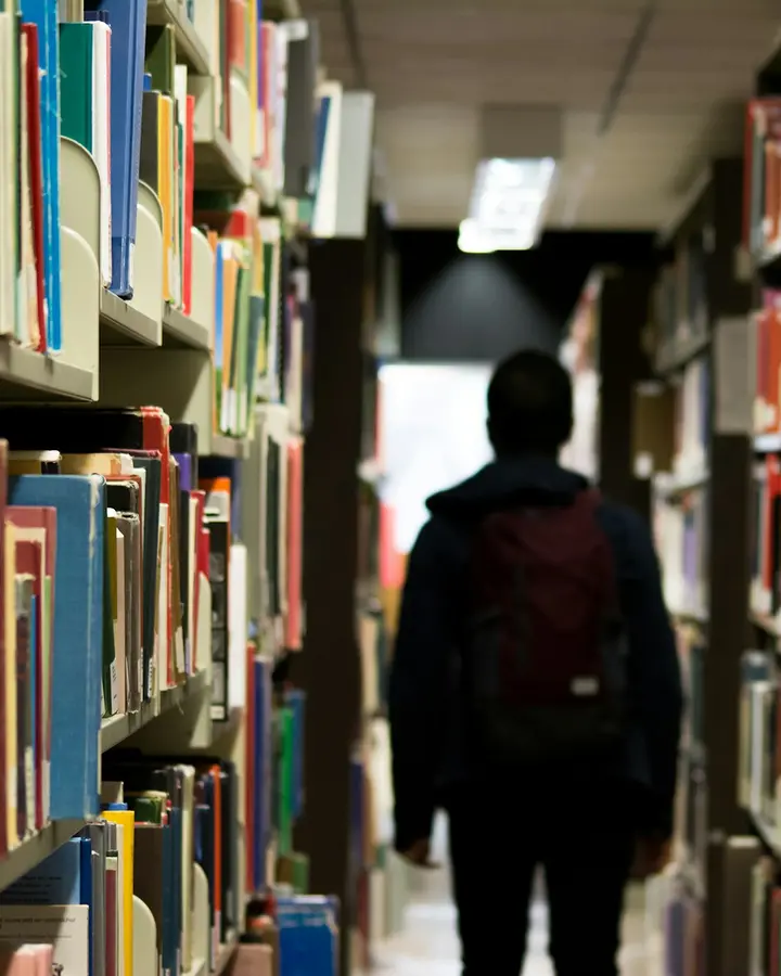 A student walking down a library aisle between tall bookshelves